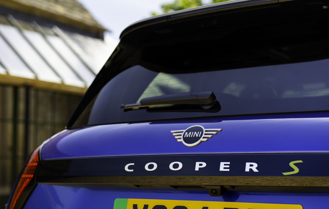Close-up of the rear end of a blue MINI Cooper Electric S, showing the MINI logo, model name, and part of a yellow license plate, with a blurred glass structure and greenery in the background.